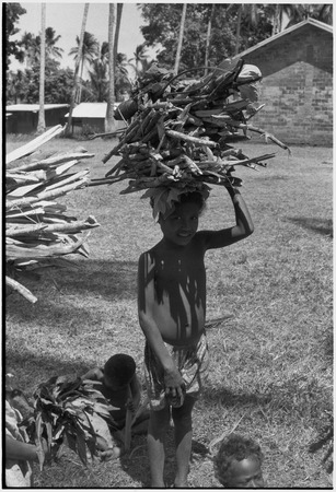 Child balances bundle of firewood on head, church in background