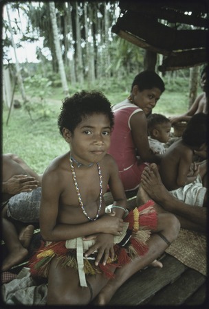 Girl wearing short fiber skirt and beaded necklace, seated under a house with other people