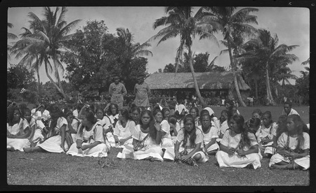 Women and men dressed in white sitting on grass, two government officials stand in the back