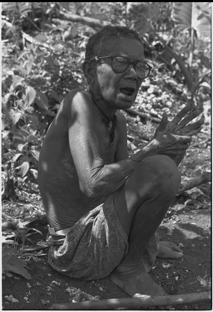 Elderly woman, Bomtavau, in a garden, she wears glasses and gestures with her hands