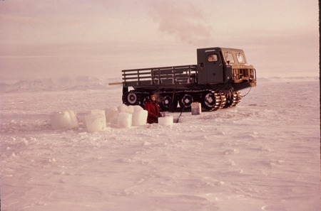 Paul Dayton cutting a hole through thick sea ice, with a Nodwell behind. near McMurdo Station, Ross Island, Antarctica. 1963