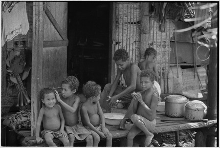 Children on a veranda, boy grates coconut into bowl, child (l) grooms younger child&#39;s hair, taro shoots (l) in background