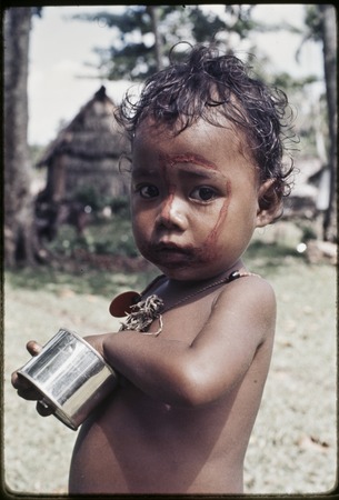 Child with betel nut paste dabbed on face and wearing a kula necklace, holds tin can