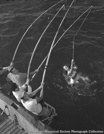 Three tuna fishermen with bamboo poles pulling in the &quot;Catch of the Year&quot; model Mary D. in a bathing suit