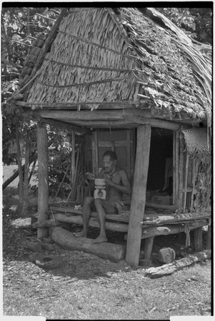 Carving: M&#39;lapokala carving a wooden bowl with a stand, probably for tourist trade, he sits on veranda of a small house