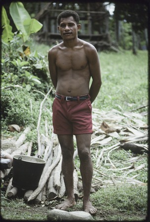 Mowaroka stands next to bucket and poles from which bark has been removed, bark (background) will be made into tapa