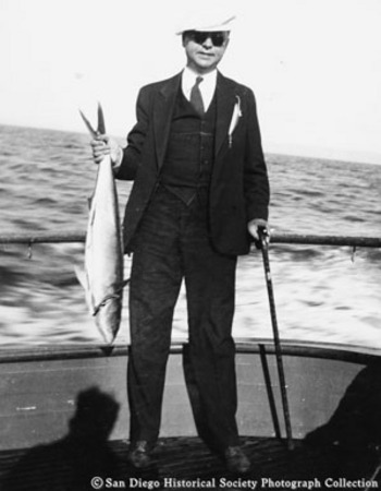 Man with cane, dressed in suit, holding fish on boat