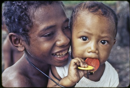 Smiling child and an infant, infant chews on a red disc commemorating Papua New Guinea&#39;s independence