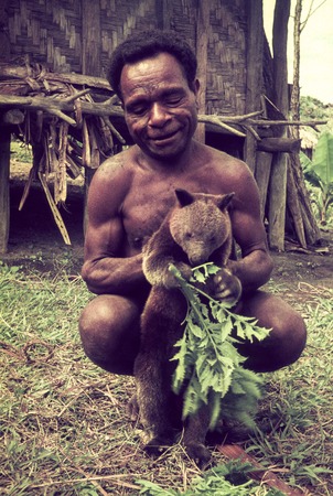 Man holding a live-caught marsupial, for later trade or consumption, house in background