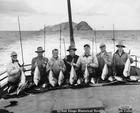 Group of sportfishermen sitting at stern of boat with fishing rods and catch of tuna, Coronado Islands in background