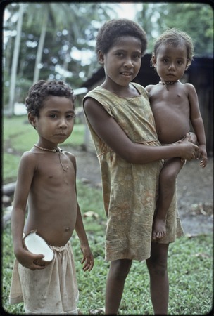 Girl holds a younger child, another girl holds split coconut, both of the younger children wear shell necklaces