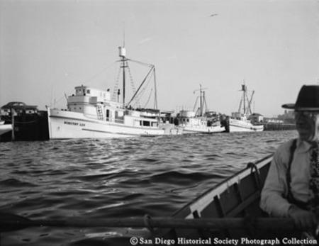View of docked tuna boats, including Dorothy Lee, with man in rowboat on right