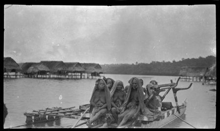 People on a double-hulled outrigger canoe, with houses on stilts over water in background