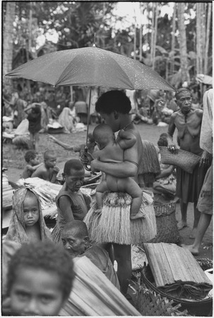 Mortuary ceremony, Omarakana: woman in short fiber skirt, holds umbrella over an infant
