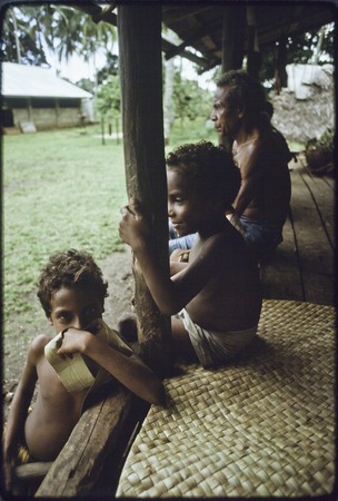 Children and man sit on veranda with pandanus mat, one child holds a dried pandanus leaf for dancing