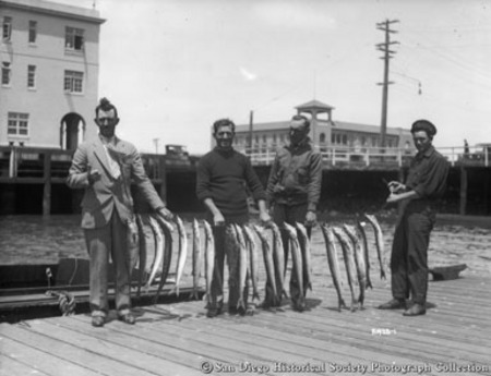 Four men on pier posing with catch of barracudas