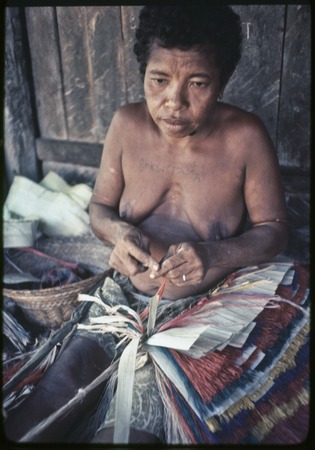 Weaving: woman makes skirt out of banana leaf fibers, some dyed red, blue or yellow, woman has word tattooed on her chest
