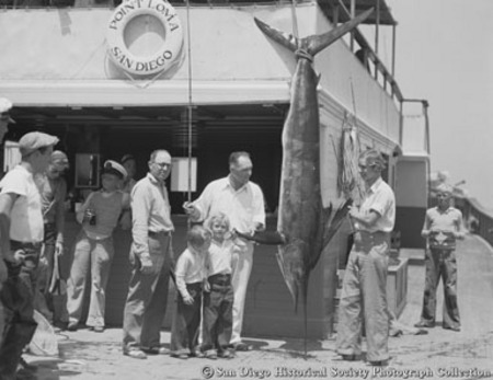 Man with fishing rod and other people standing around marlin on display, Point Loma
