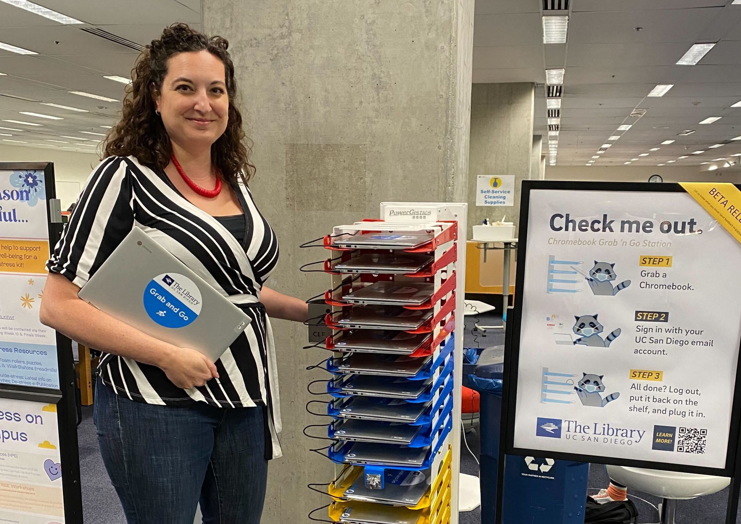 Allison Flick, manager of Service Operations for the UC San Diego Library, holding a Chromebook and standing next to a Chromebook loan station in Geisel Library.