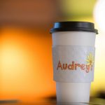 Coffee cup with branded Audrey's sleeve sitting on counter with back lighting