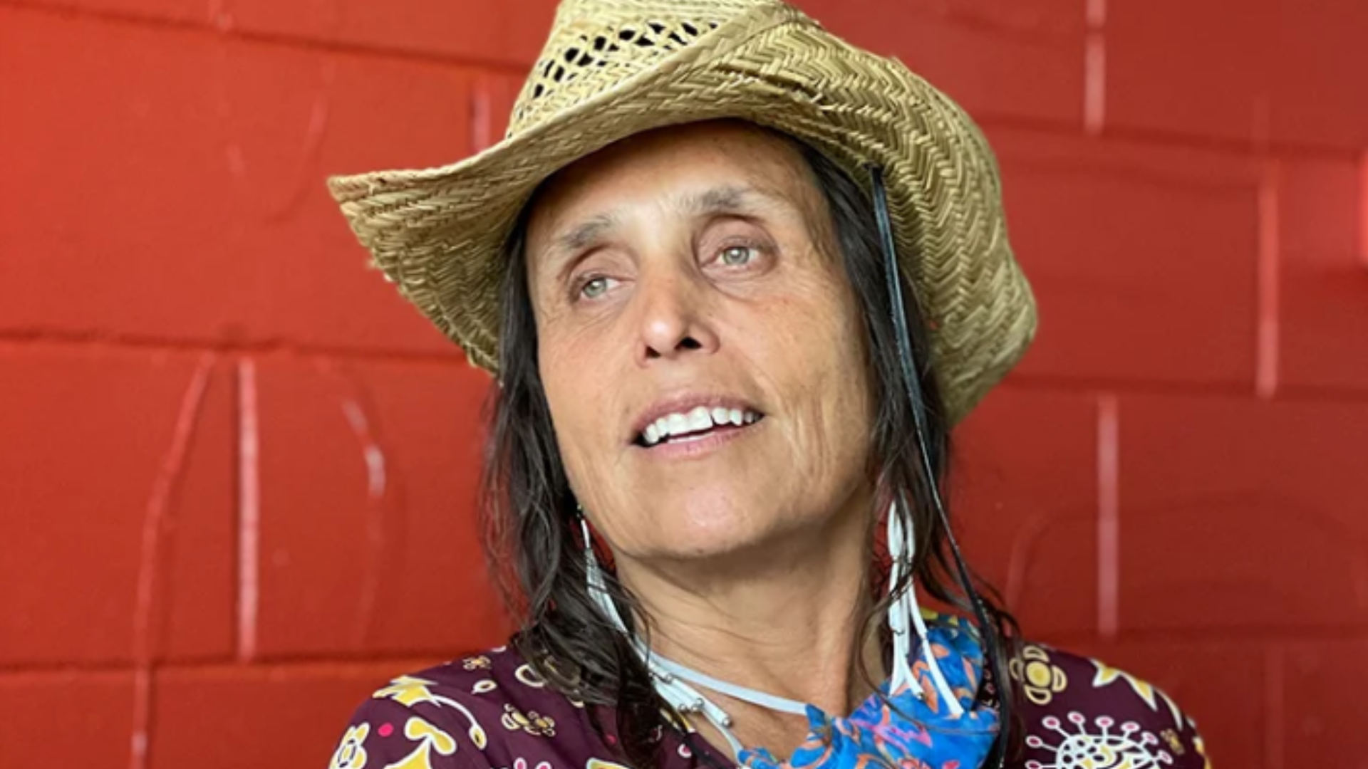 Phot of Winona LaDuke in front of a red brick wall wearing a straw hat.