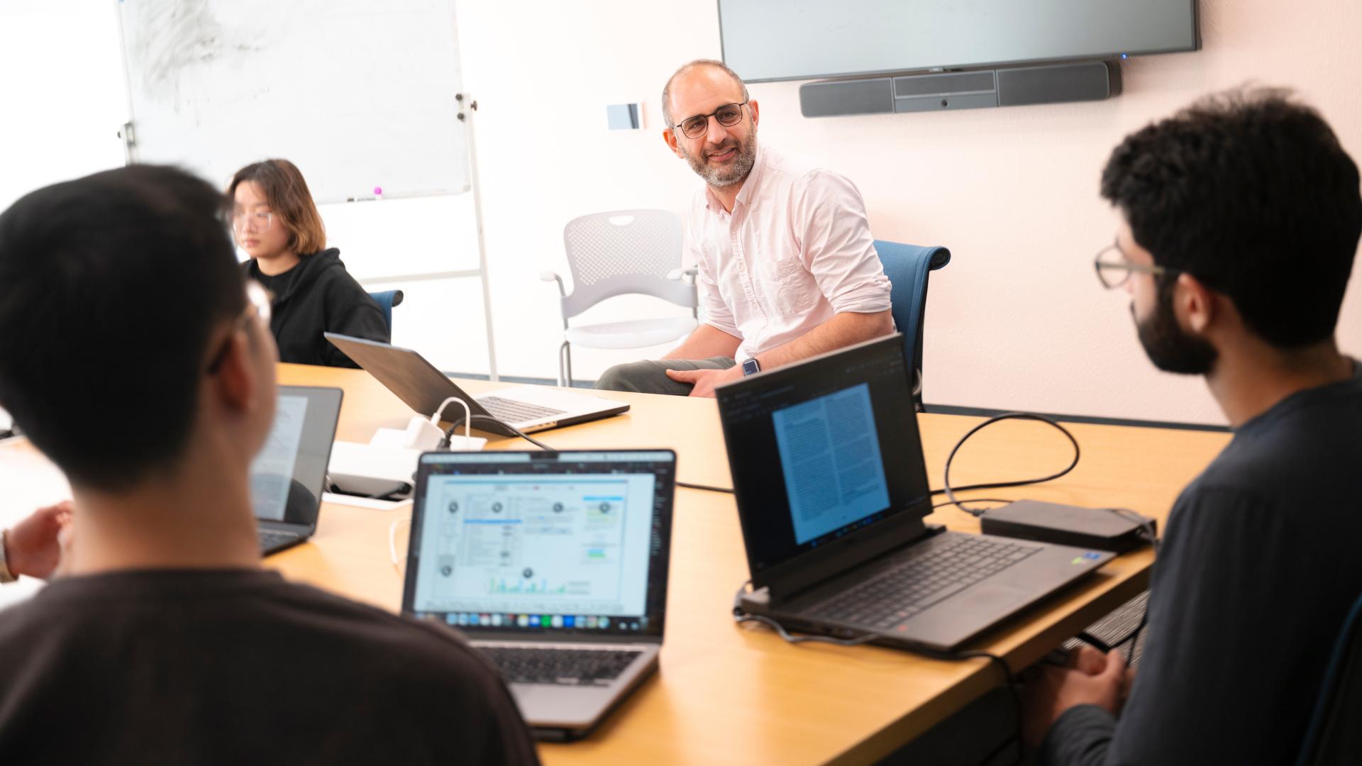 Students with laptops sitting at a tabling talking to a professor